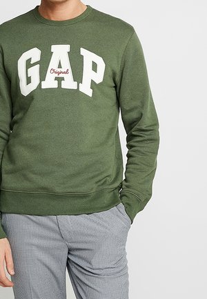 Mężczyzna noszący oliwkową bluzę GAP z napisem „Original”, w połączeniu z czarno-białymi, kratowanymi spodniami, ręką w kieszeni.