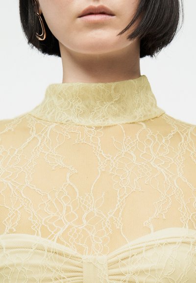 Γυναίκα με κοντά σκούρα μαλλιά που φορά ένα διάφανο μπεζ δαντελένιο τοπ με ψηλό λαιμό και ένα χρυσό σκουλαρίκι κρίκο, σε λευκό φόντο.