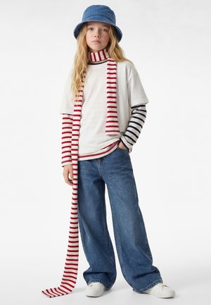 Jongere in wijde jeans, witte sneakers, gelaagde gestreepte lange mouwen, wit T-shirt, rood-wit gestreepte sjaal en blauwe bucket hat.