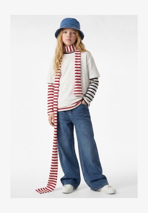 Joven con vaqueros de pierna ancha, zapatillas blancas, camiseta de manga larga de rayas en capas, camiseta blanca, bufanda de rayas rojas y blancas, y sombrero tipo cubo azul.