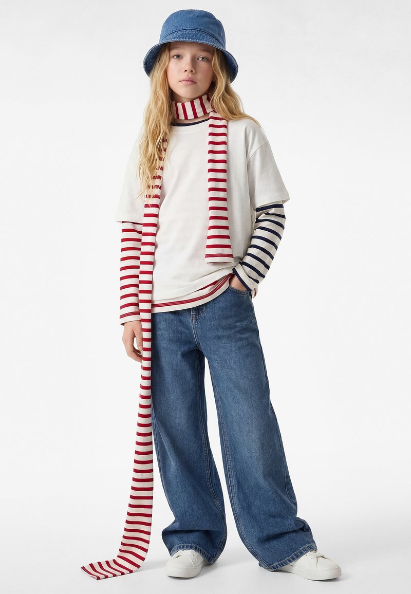 Persona giovane con jeans a gamba larga, sneakers bianche, maglia a maniche lunghe a righe sovrapposte, t-shirt bianca, sciarpa a righe rosse e bianche, e cappello a secchiello blu.