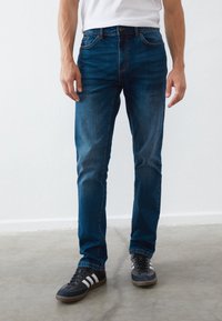 Dark blue denim jeans with a classic cut, featuring five pockets, and subtle fading. Paired with black and white athletic shoes.