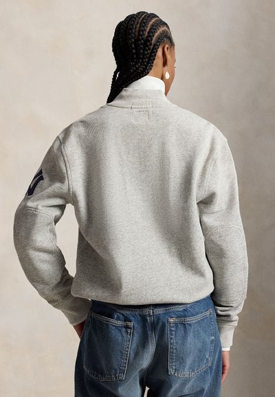 Personne aux cheveux tressés portant un sweat gris avec un patch bleu marine sur la manche et un jean bleu, vue de dos sur un fond uni.