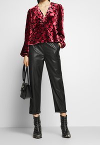 Blusa in velluto rosso con motivo floreale, pantaloni in ecopelle neri e stivaletti neri con borchie. La modella tiene una piccola borsa a tracolla nera.