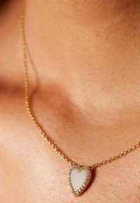 Gouden ketting met een hartvormige hanger, met een lichte steen in het midden omringd door kleine heldere edelstenen. Gladde, gepolijste afwerking.