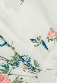 Witte stof met een bloemenpatroon met blauwe en roze bloemen, verweven groene bladeren en een geknoopt detail in het midden.