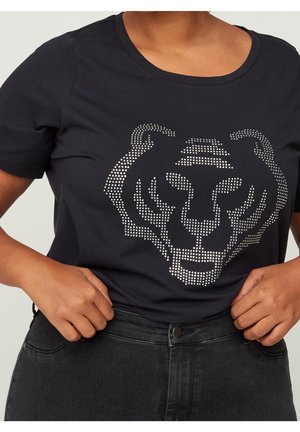 Personne portant un t-shirt noir orné d'un motif en strass représentant un visage de tigre, associé à un jean noir taille haute.