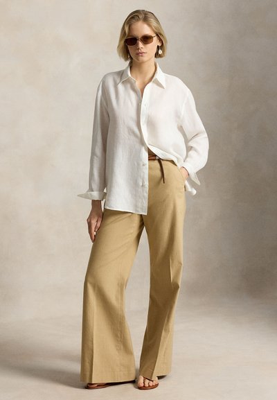Ξανθιά γυναίκα που φορά καφέ γυαλιά ηλίου, λευκό πουκάμισο με κουμπιά, μπεζ παντελόνι με φαρδύ πόδι, σανδάλια και λευκά σκουλαρίκια, στέκεται με το ένα χέρι στην τσέπη.