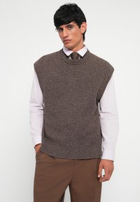 Brauner, gestrickter ärmelloser Pullover mit geripptem Muster, Rundhalsausschnitt, kombiniert mit einem hellfarbigen Hemd und einer gemusterten Krawatte, getragen mit braunen Hosen.