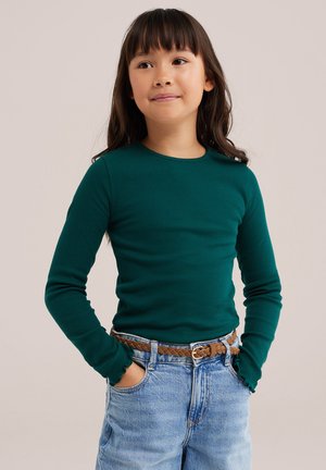 Une jeune fille pose, les mains dans les poches de son jean. Elle porte un haut vert foncé et une ceinture marron tressée, sur un fond uni.