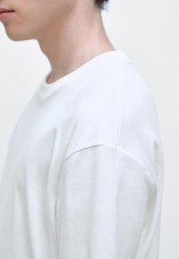 White cotton long-sleeve shirt with a round neckline and subtle shoulder seams, displaying a smooth texture and a fitted silhouette.
