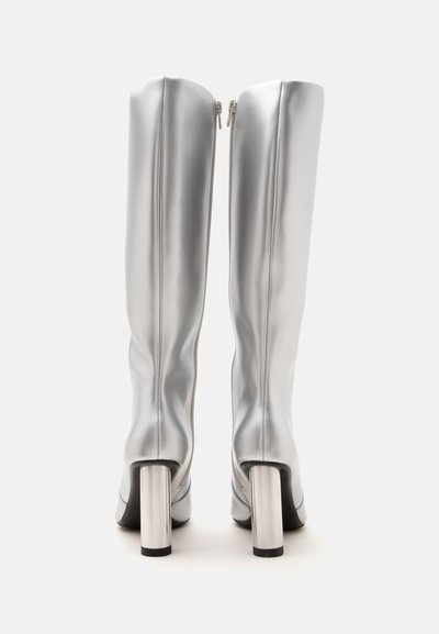 Paire de bottes argentées brillantes montant jusqu'aux genoux avec talons carrés, fermeture éclair à l'arrière, debout sur un fond blanc.