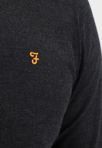 Czarna tkanina o teksturowanym wykończeniu, z małym pomarańczowym haftowanym logo na lewym ramieniu. Minimalistyczny design i lekkość.