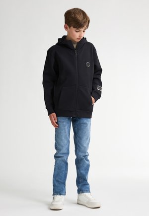 Hoodie noir à zip avec poches avant et logo, associé à un jean bleu clair et des baskets blanches. Tissu lisse et coupe décontractée.