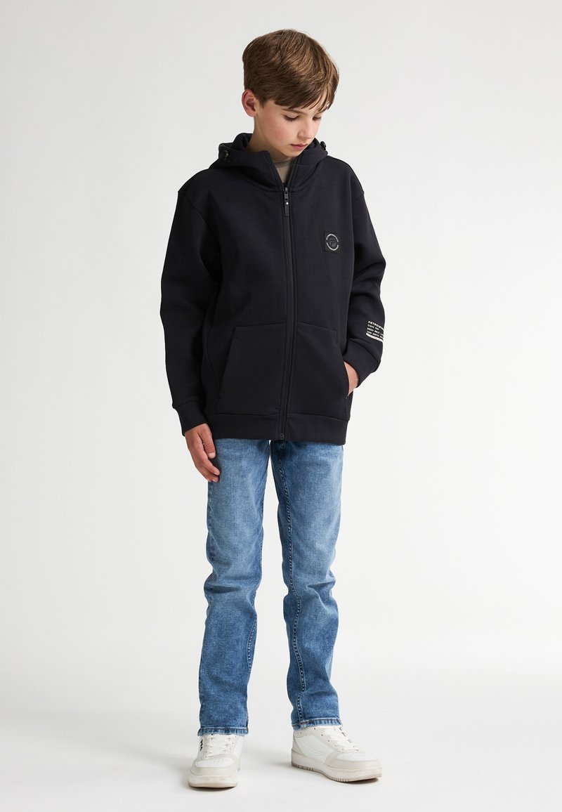 Sudadera negra con cremallera, bolsillos frontales y un logo, combinada con jeans azul claro y zapatillas blancas. Tejido suave y corte relajado.