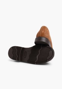 Rudo suede šlepetė su apvalia nosimi, siuvinėjimo detalėmis ir žemu juodu guminės pakulne. Textūruota juoda padas su banguotu raštu.