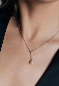 Srebrny naszyjnik z delikatnym łańcuszkiem, z pojedynczą okrągłą perłą zawieszoną na małym akcentującym pomponiku. Gładka tekstura.