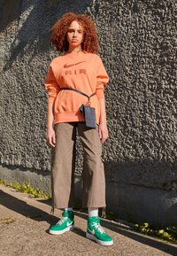 Orange sweatshirt med "AIR" text, beige cargobyxor och gröna högstövel-sneakers. Inkluderar en liten svart påse fäst med ett bälte.