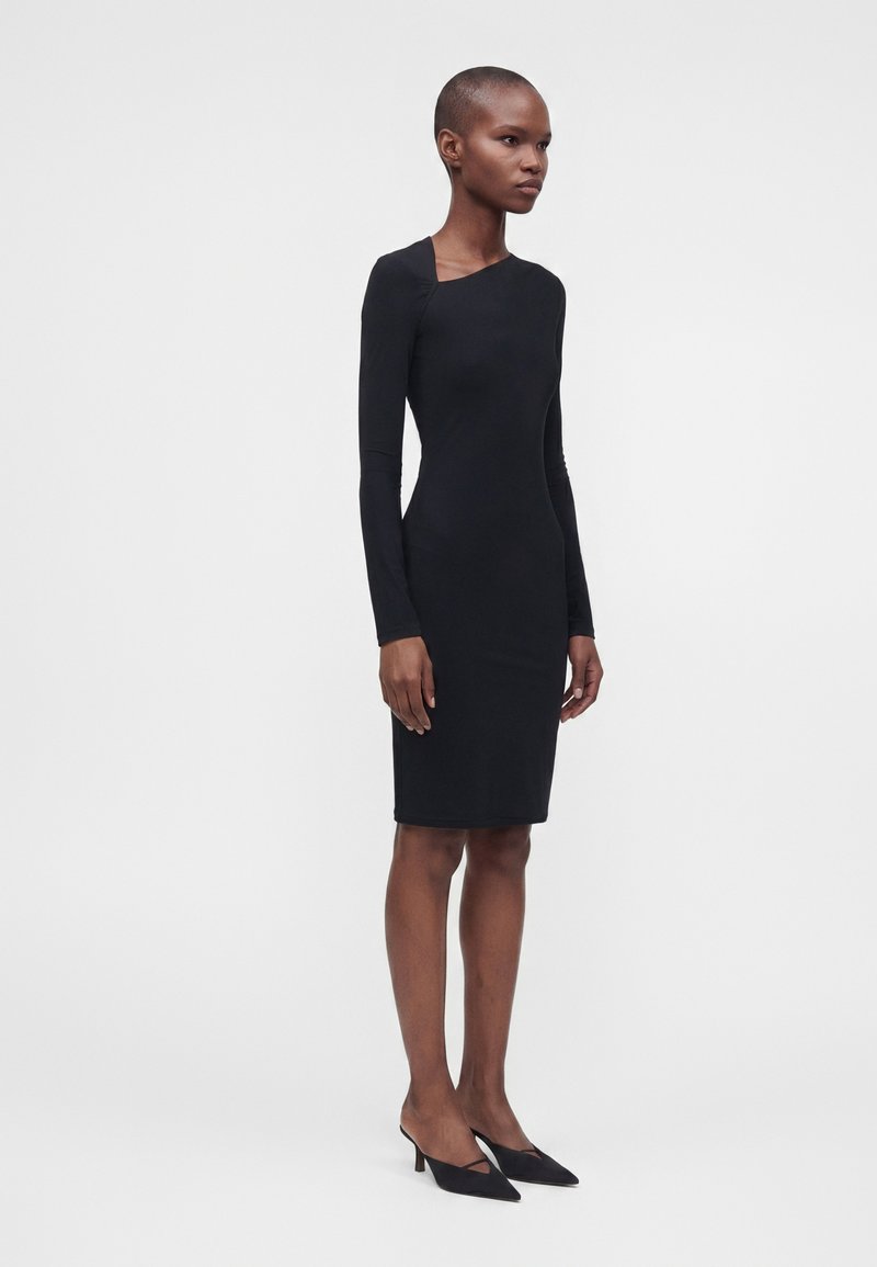 Femme debout, portant une robe noire près du corps à manches longues et longueur genou, ainsi que des escarpins noirs à bout pointu, sur un fond clair uni.