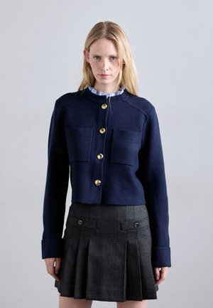 Giacca a maglia blu navy con bottoni dorati, due tasche sul petto e un design aderente. Abbinata a una gonna plissettata a quadri con bottoni frontali.