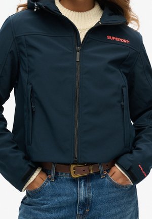 Personne portant une veste zippée Superdry bleu marine sur un pull en maille crème, les mains dans les poches, associée à un jean bleu et une ceinture marron.
