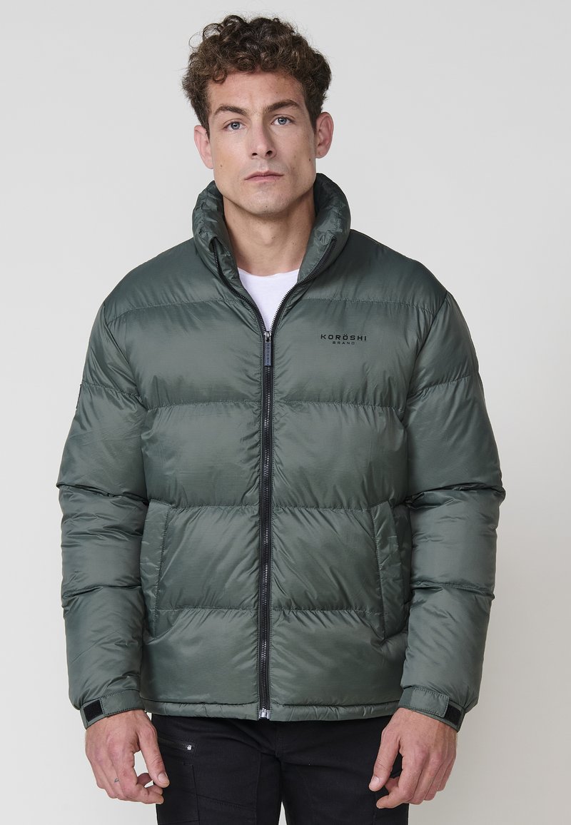 Chaqueta acolchada de color verde oscuro con un alto cuello, cierre frontal con cremallera y diseño de costuras horizontales. Presenta un logo en el pecho y puños elásticos.