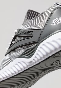 Zapatilla deportiva gris con parte superior de punto, cuello acanalado y suela de goma con patrones texturizados. Logo visible en el lado y en los cordones.