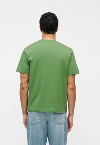 Άνδρας με σγουρά σκούρα μαλλιά που φοράει απλό πράσινο κοντομάνικο μπλουζάκι και ανοιχτόχρωμα μπλε τζιν, γυρισμένος με την πλάτη σε λευκό φόντο.