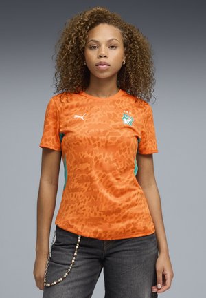 Frau mit lockigem Haar, die ein orangefarbenes Puma-Fußballtrikot mit einem grünen und weißen Abzeichen trägt, kombiniert mit dunklen Jeans und einer Perlenketten-Accessoire.