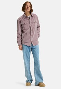 Lichtpaarse corduroy shirt met twee borstzakken, knoopsluiting en bruine knopen. Gecombineerd met lichtblauwe wijde jeans en beige schoenen.