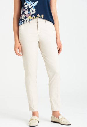 Frau trägt cremefarbene Slim-Fit-Hose, marineblaues Oberteil mit Blumenmuster und cremefarbene Loafer mit goldener Kettendetail, steht vor weißem Hintergrund.