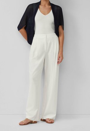 Vrouw die witte, hoge taille broek met wijde pijpen draagt, een wit V-hals topje, een marineblauwe korte mouwen gebreide shrug en beige platte sandalen, staand tegen een grijze muur.