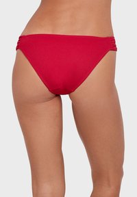 Bas de bikini rouge avec texture côtelée, accents gather sur les côtés, et un design taille basse, avec une finition lisse et une coupe ajustée.