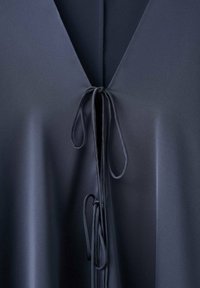 Blouse bleu marine avec un col en V, dotée de fermetures à nouer au niveau de l'encolure. Le tissu semble lisse, léger et doté d'une texture douce.