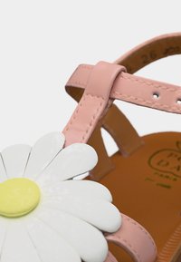 Bride de sandale rose avec des détails cousus et une grande décoration de marguerite blanche et jaune sur une semelle beige.