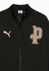Veste noire zippée avec col côtelé, logo Puma beige sur la poitrine gauche et patch beige texturé en forme de lettre "p" sur la poitrine droite.