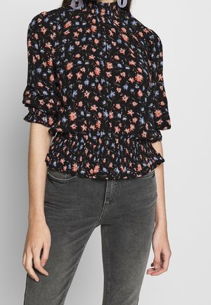 Vrouw draagt een zwarte bloemenblouse met geplooide taille en mouwen, gecombineerd met vervaagde zwarte spijkerbroek tegen een effen achtergrond.