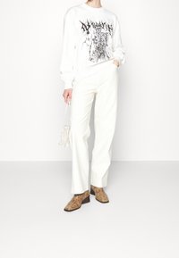 Vit sweatshirt med svart grafisk design, matchad med vita rakbeniga byxor och bruna stövletter i ormskinnsprint. Liten vävd vit väska.