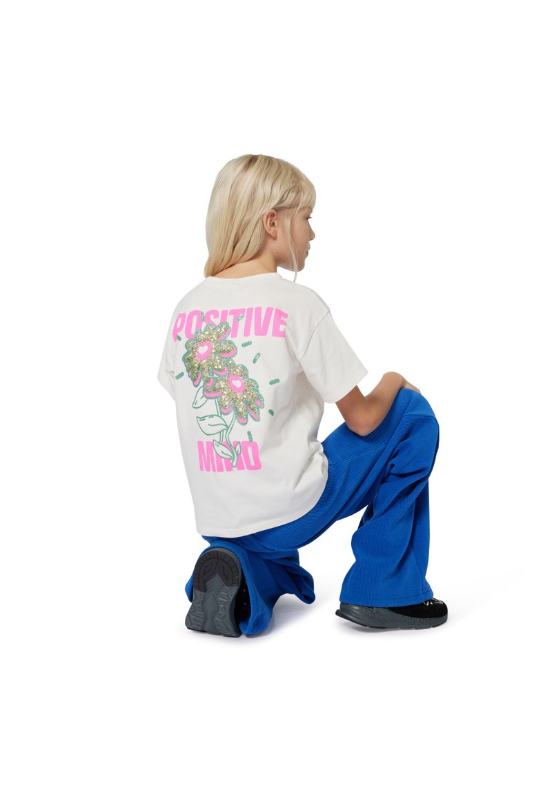 Kind met kort blond haar knielt en draagt een wit T-shirt met "Positive Mind" en een bloemenontwerp, gecombineerd met een blauwe broek en zwarte schoenen.