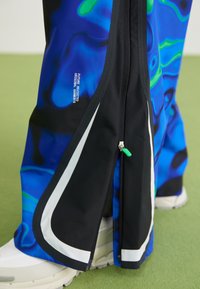 Nærbillede af blå og sorte bukser med en grønaccentueret lynlås og hvide sneakers på grønne gulv, med teksten "FUTURE REVISITED ORIGINAL GARMENTS".
