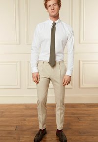 Hellbeige, maßgeschneiderte Hose mit flachem Bund, kombiniert mit einem weißen Hemd und einer olivgrünen Krawatte. Braune Slipper und kastanienrote Socken.