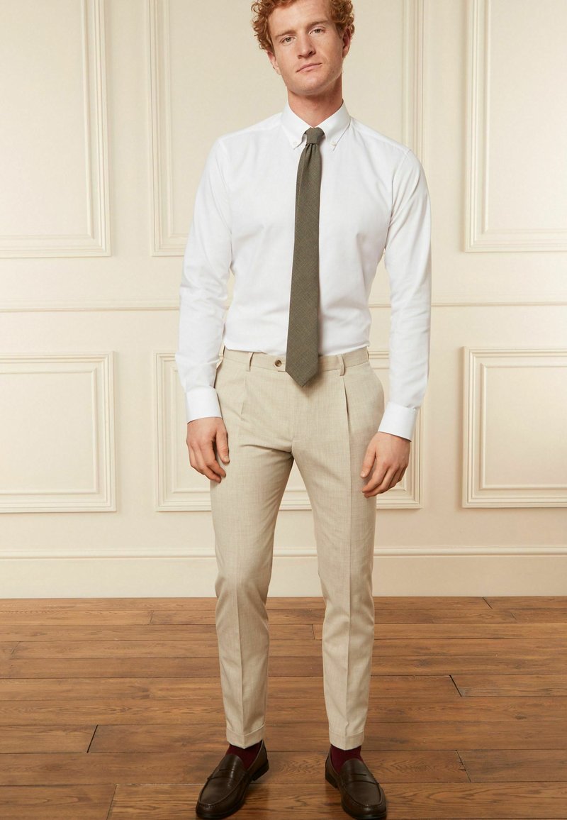 Hellbeige, maßgeschneiderte Hose mit flachem Bund, kombiniert mit einem weißen Hemd und einer olivgrünen Krawatte. Braune Slipper und kastanienrote Socken.