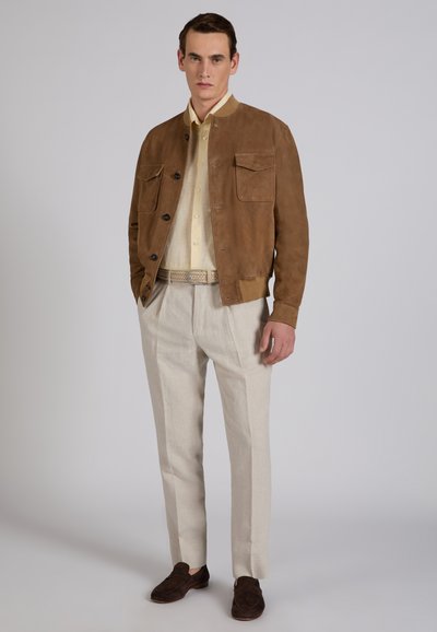 Man met een bruine suède jas over een beige overhemd, lichte beige broek, gevlochten riem en donkerbruine loafers, staand tegen een effen achtergrond.