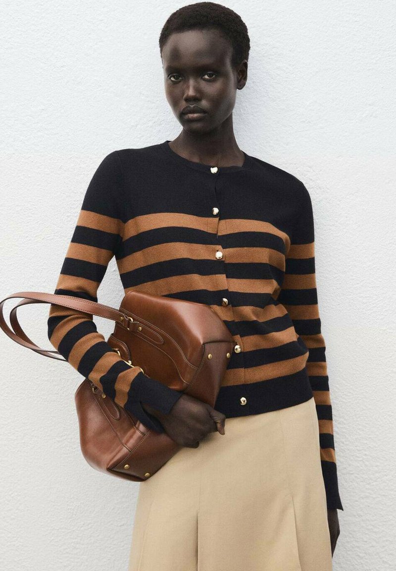 Marinblauer und braun gestreifter Cardigan mit goldenen Knöpfen und langen Ärmeln. Das Model hält eine braune Ledertasche. Ein schlichter beiger Rock rundet den Look ab.