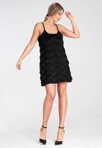 Schwarzes Kleid mit schichtiger Fransenstruktur, verstellbaren Spaghettiträgern und einer taillierten Silhouette, kombiniert mit schwarzen High Heels.