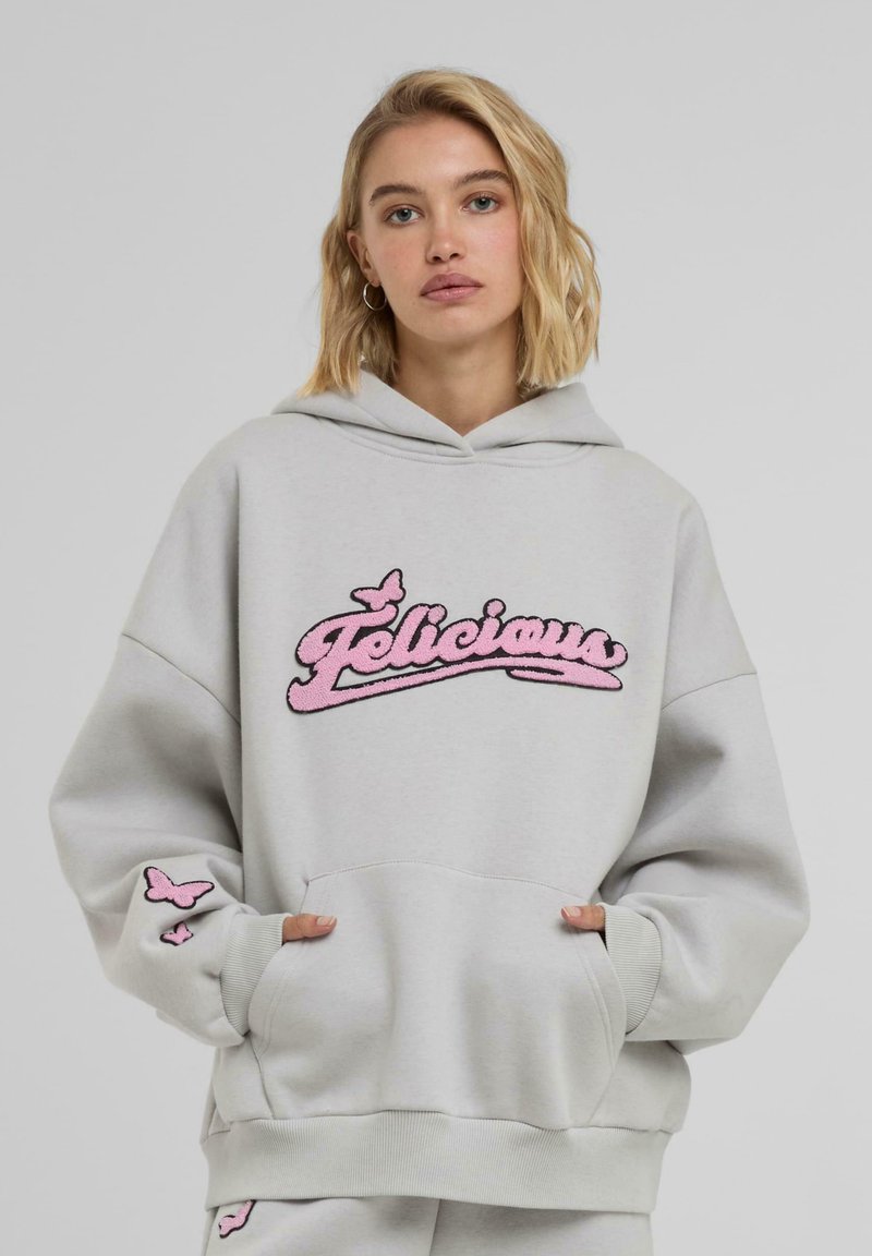 Sivi dukserica s labavim krojem, sa ružičastim izvezenim logotipom "Felicious" i malim leptirima na rukavima i džepu.