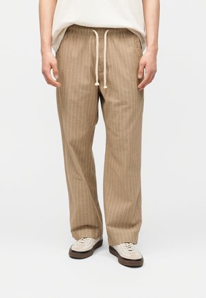 Férfi, bézs csíkos, húzózsinóros nadrágot és bézs tornacipőt viselve, egyszínű világos háttér előtt áll.