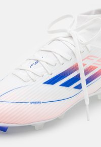 Vita fotbollsskon med blå och röda ränder, vita snören knutna i en båge, och texten "SPRINTGRID" på sidan.