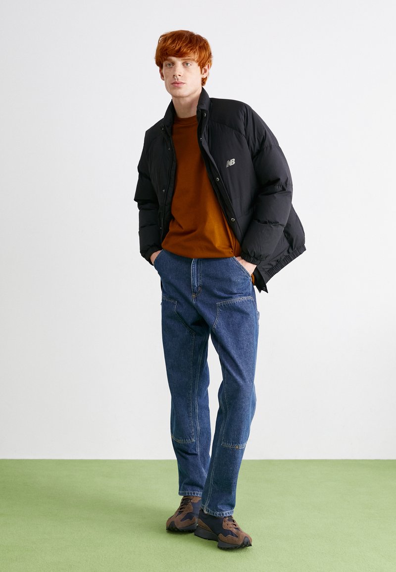 Schwarze Steppjacke mit hohem Kragen, brauner Sweatshirt, blaue weit geschnittene Jeans und braune Sneaker mit strukturierten Details. Schlichter Hintergrund.