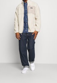 Hombre con una chaqueta de forro polar blanca sobre una camisa de botones azul, jeans oscuros arremangados y zapatillas blancas, de pie frente a un fondo liso.